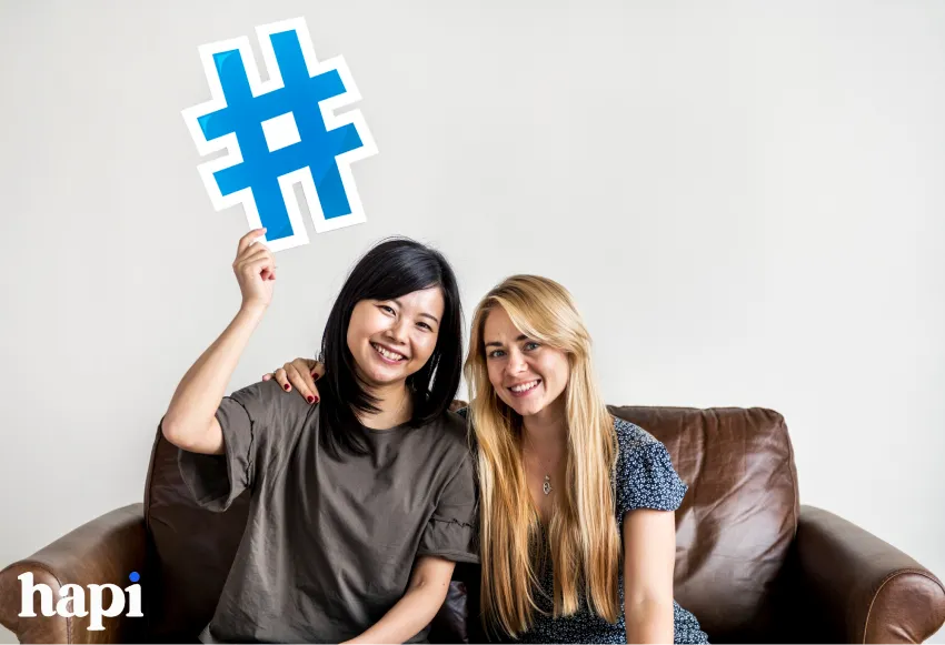 Two women sitting on a couch holding a Hashtag symbol cutout